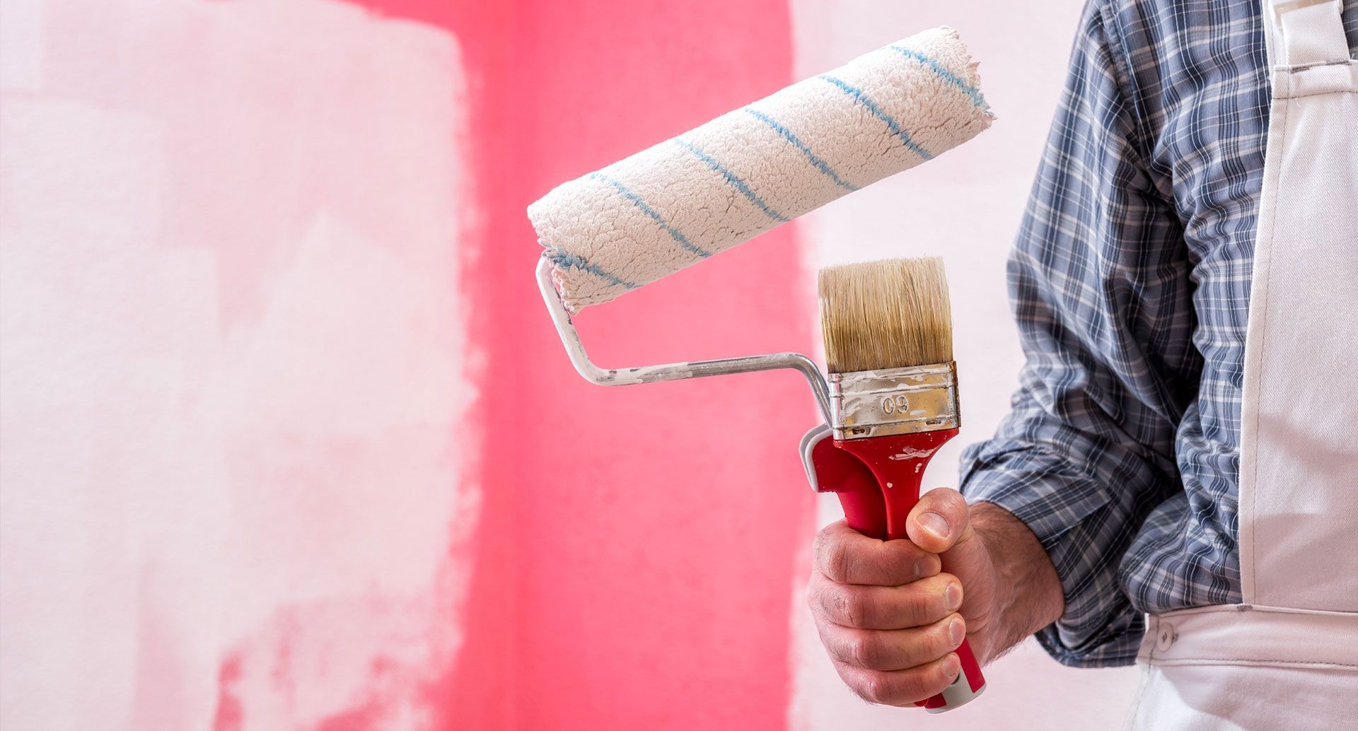 Una persona sostiene una herramienta combinada de rodillo y brocha frente a una pared parcialmente pintada de rosa.