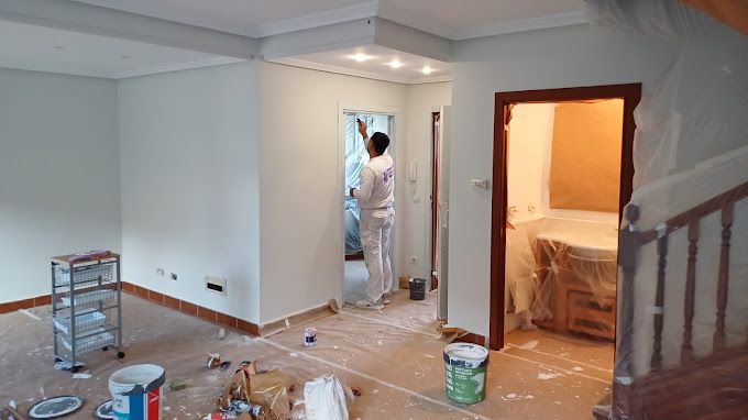 Una persona vestida con ropa de trabajo blanca pinta el marco de una puerta en una habitación en obras.