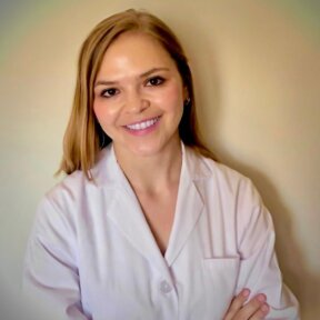 Una mujer con una bata de laboratorio blanca sonríe con los brazos cruzados.