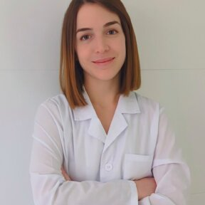 Mujer con bata blanca de laboratorio con los brazos cruzados, sonriendo.