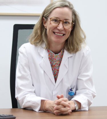 Una mujer que lleva gafas, una bata blanca y una blusa estampada sonríe y se sienta en un escritorio.