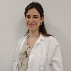 Una mujer con bata blanca sonríe, en el interior, frente a una pared blanca.