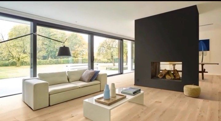 Sala de estar moderna con grandes ventanales, sofá de color claro, pared de chimenea oscura y suelo de madera clara.