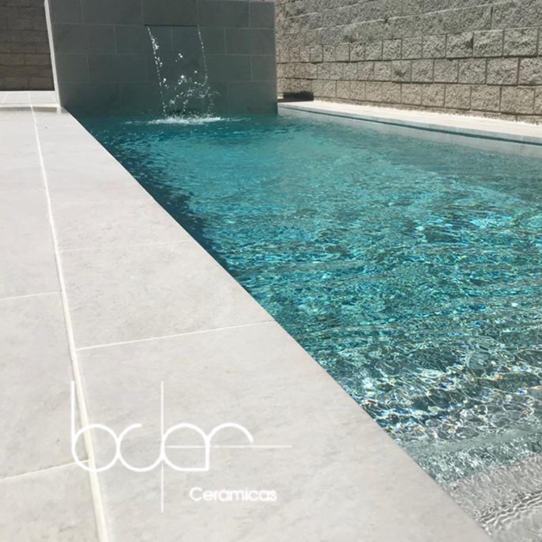 Piscina exterior moderna con agua turquesa, terraza de azulejos y cascada de pared de piedra.