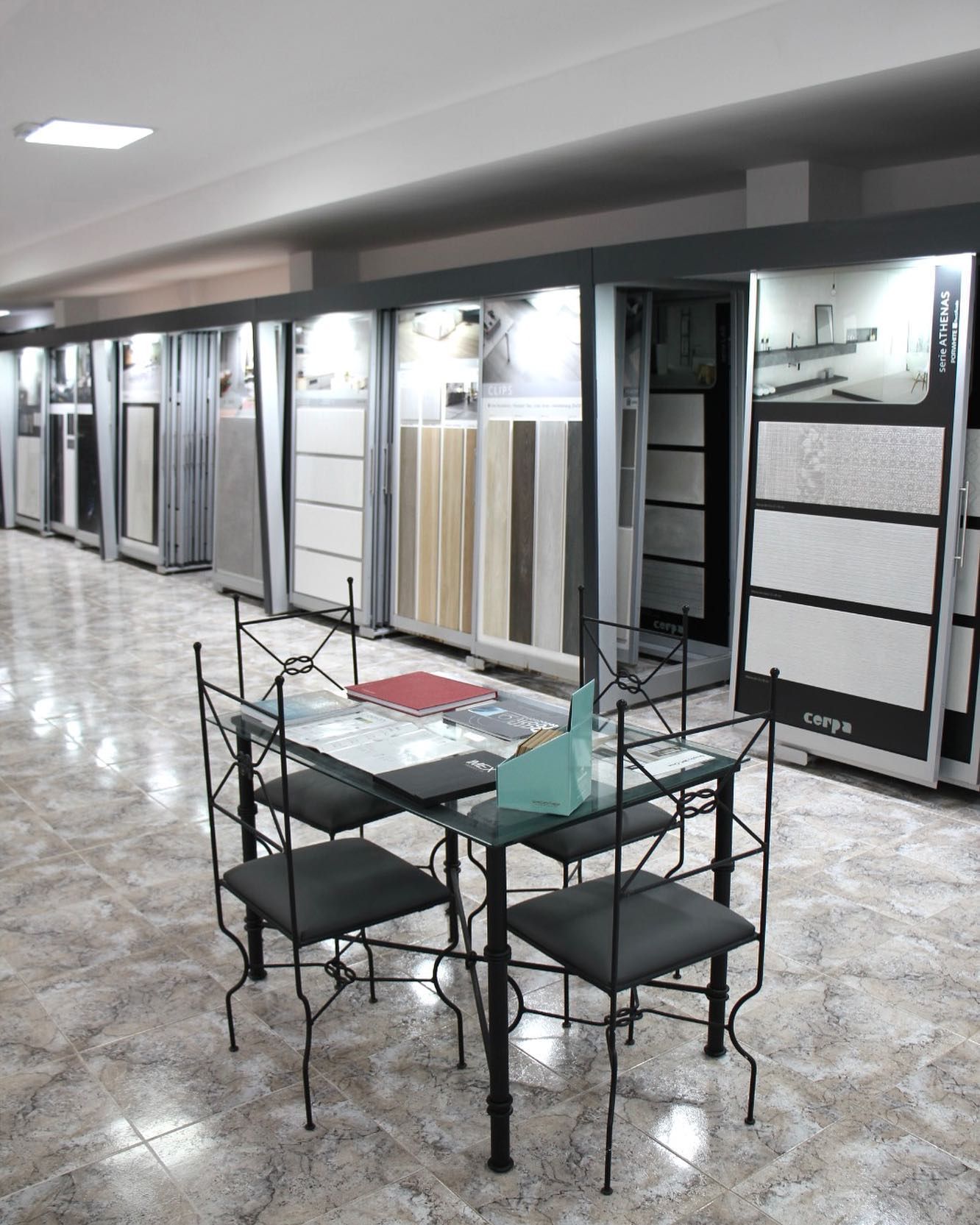 Pasillo vacío con azulejos, habitaciones con fachada de cristal y una pequeña mesa y sillas en primer plano.