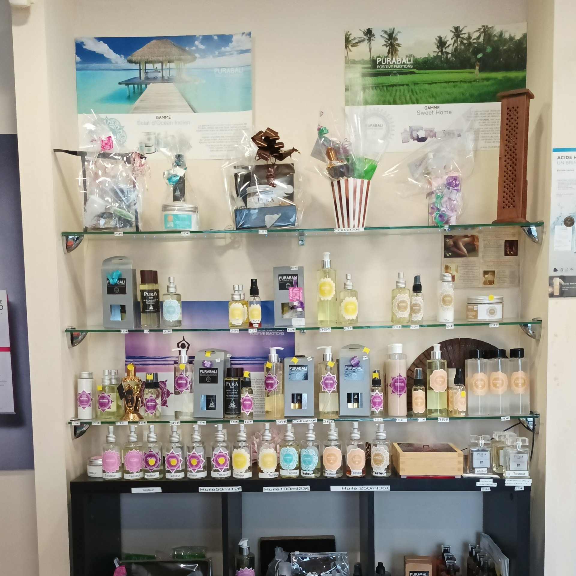 Étagères en verre exposant une large gamme de produits de bien‑être et de parfums, dont des sprays, huiles et coffrets cadeaux, avec deux affiches décoratives au mur en arrière‑plan.