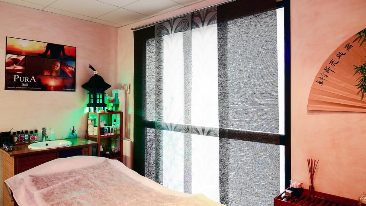 Salle de soins zen avec table de massage, éclairage doux, étagère de produits bien‑être et décor asiatique.