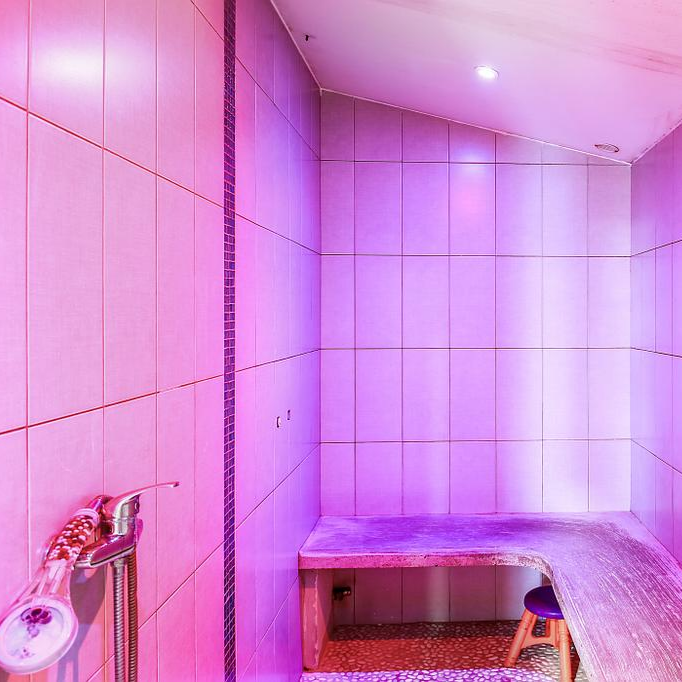 Pièce de hammam carrelée éclairée en rose, avec un banc en pierre et un jet de douche mural.