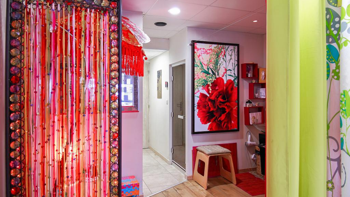 Entrée colorée d’un centre de bien‑être, décorée de rideaux de perles rouges, d'un grand tableau floral et d’étagères murales.