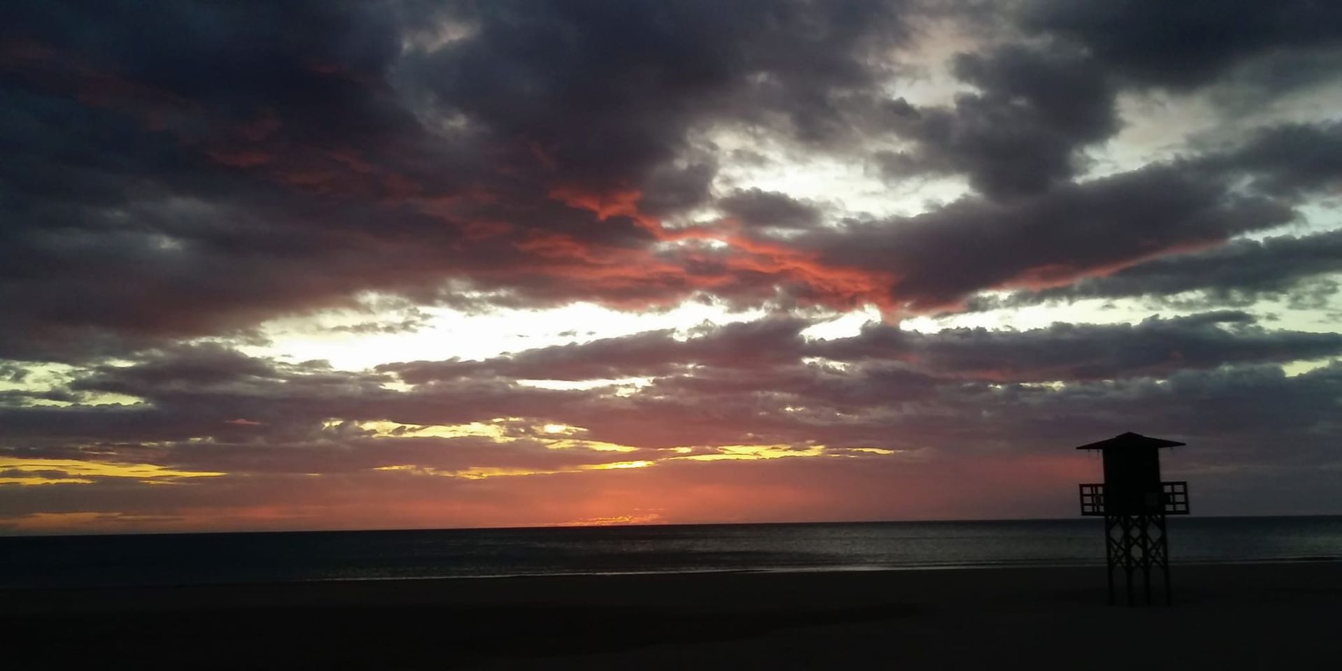 Puesta de sol sobre una costa oscura con nubes dramáticas y una torre de vigilancia recortada contra el horizonte a la derecha.