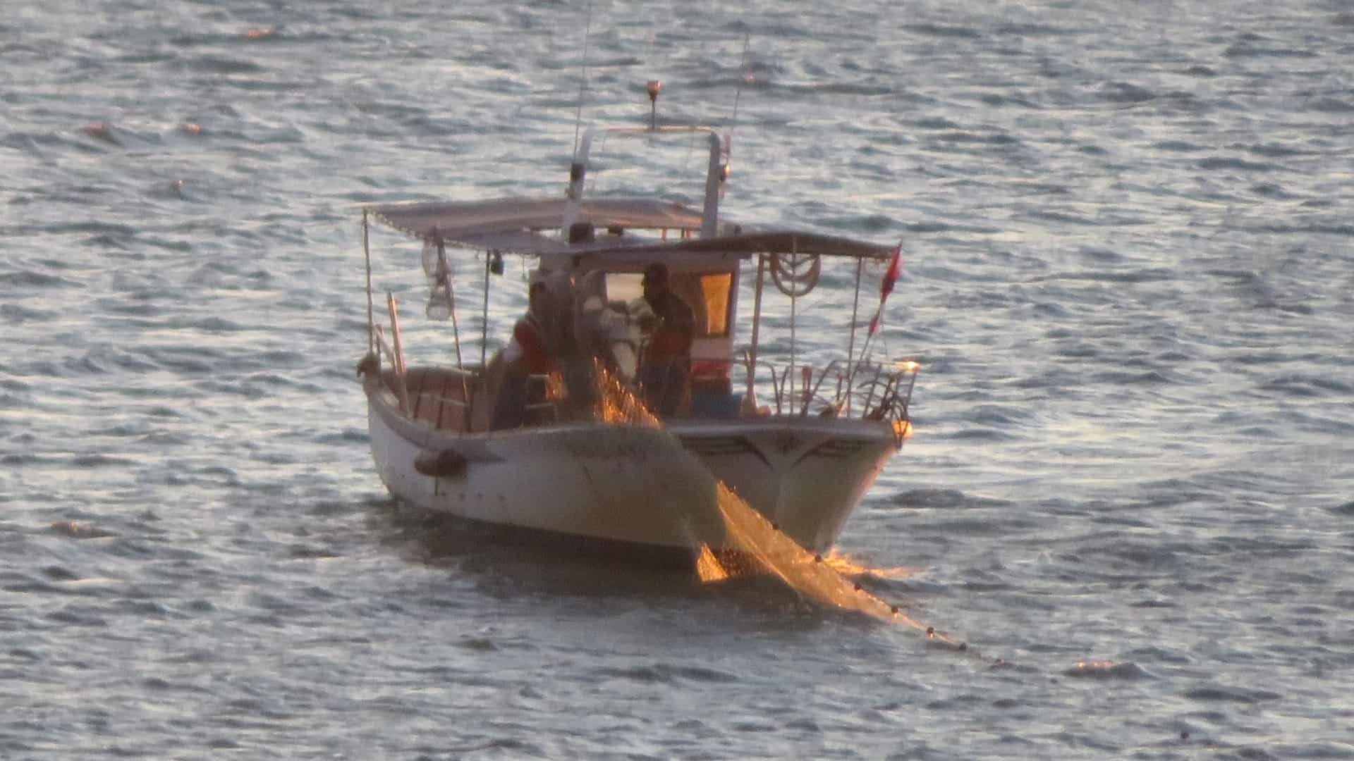 Pequeña embarcación en aguas abiertas con una hoguera encendida en la proa, vista desde la distancia.