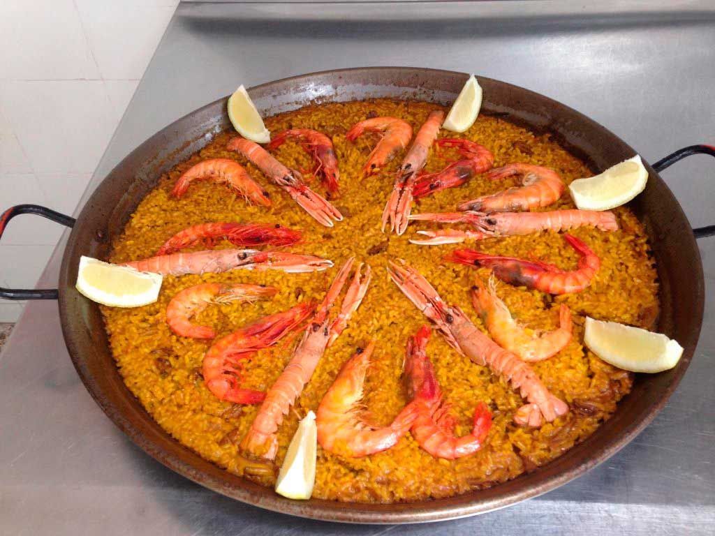 Gran paellera con gambas, salchicha, arroz y rodajas de limón dispuestas encima.