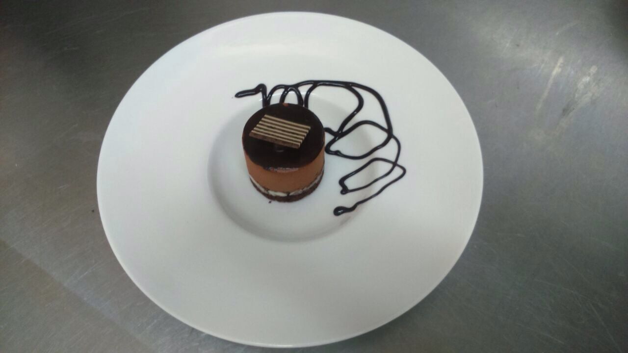 Pequeño postre de chocolate con glaseado oscuro sobre un plato blanco en una superficie de acero inoxidable.