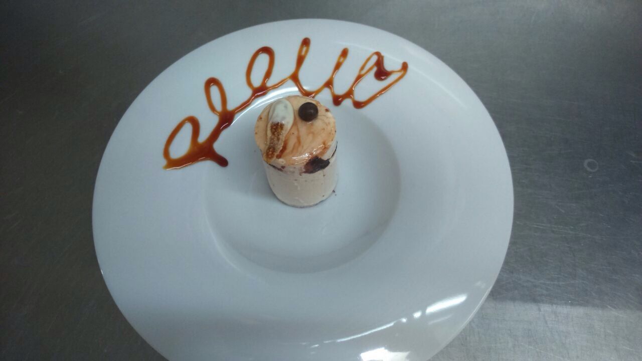 Plato blanco con un pequeño postre cilíndrico cubierto con crema batida y salsa de chocolate, con la inscripción encima.