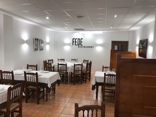Comedor vacío de un restaurante con paredes blancas, sillas de madera y manteles a cuadros bajo luces brillantes del techo.