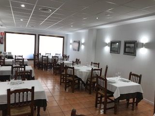 Comedor vacío de un restaurante con manteles blancos, sillas de madera, suelo de baldosas y ventanas luminosas.