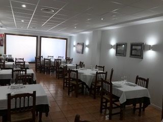 Comedor vacío de un restaurante con manteles blancos, sillas de madera, suelo de baldosas y ventanas luminosas.