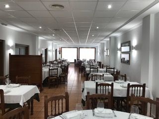 Comedor vacío de un restaurante con manteles blancos, sillas de madera y ventanas luminosas al fondo.