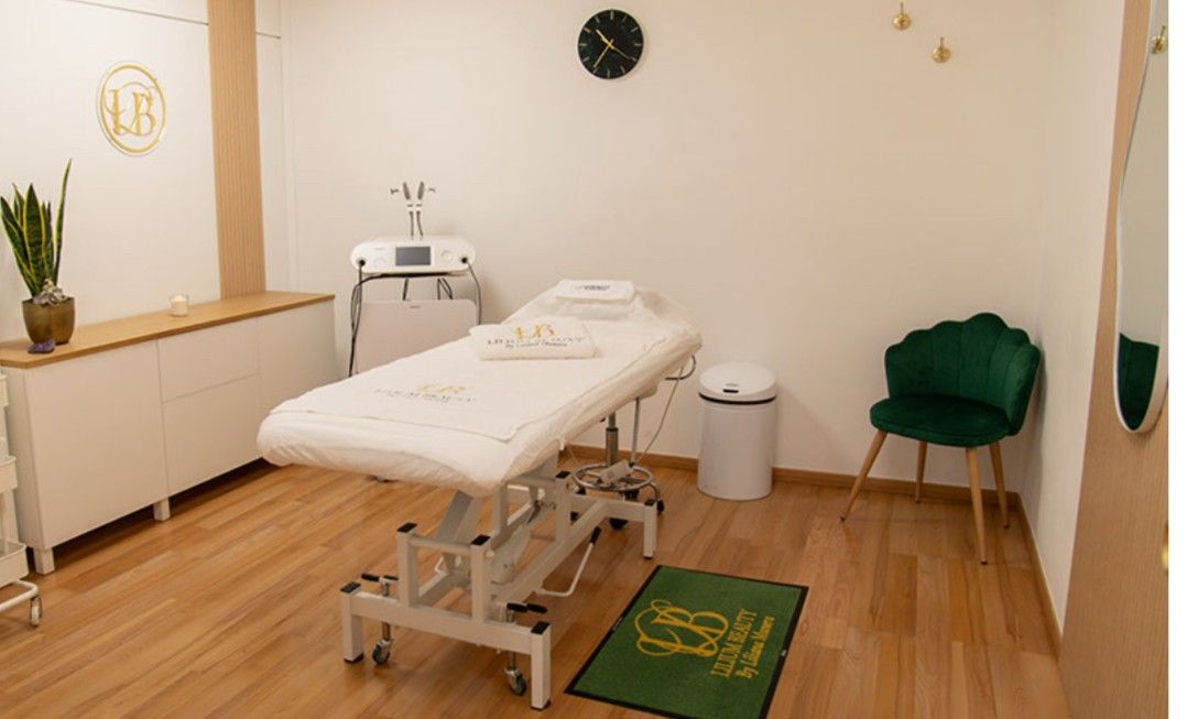 Sala de tratamientos con cama blanca, sillón verde y equipo médico. Suelos de madera y paredes blancas.