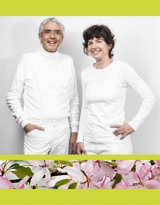 Zwei Personen in passenden weißen Langarmhemden lächeln vor einem weißen Hintergrund über einem Streifen rosa Blumen.
