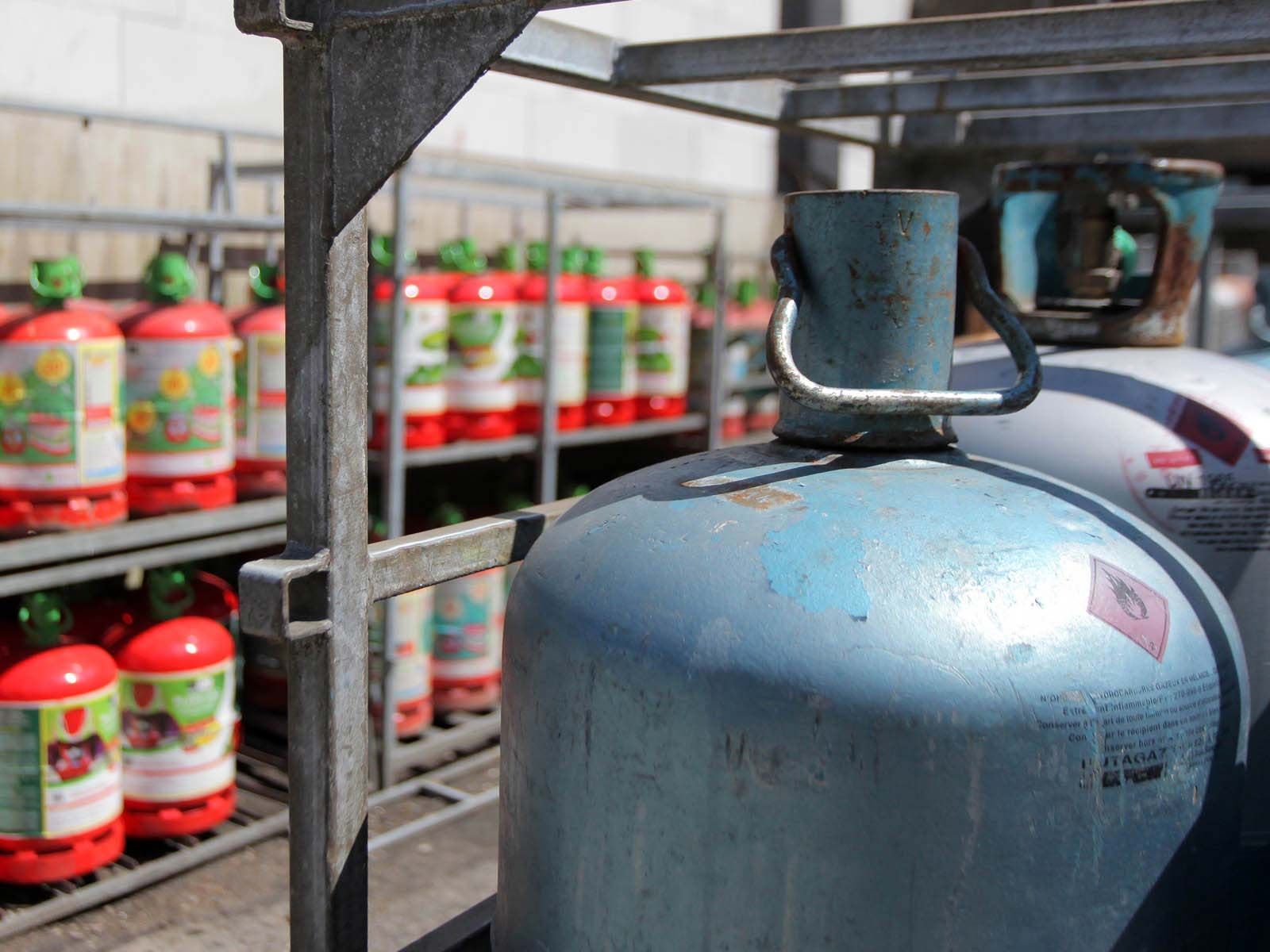 Au premier plan, une bonbonne de propane bleu clair est posée, derrière laquelle sont rangées des rangées de bouteilles de gaz rouges rangées sur des étagères.