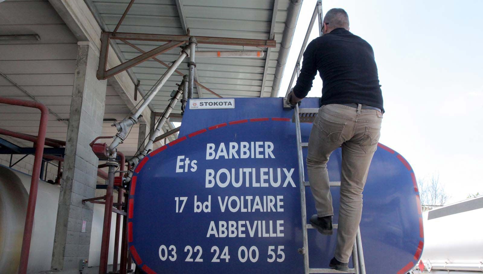 Une personne grimpant à une échelle à l'arrière d'un camion-citerne bleu portant l'inscription Ets Barbier Boutleux et des informations de contact.