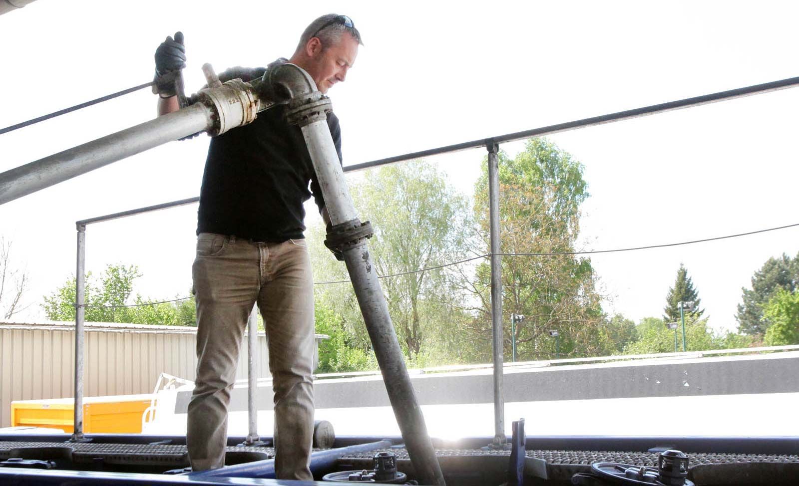 Une personne en tenue de travail utilise un tuyau pour pomper une substance dans une grande cuve industrielle d'une installation extérieure.