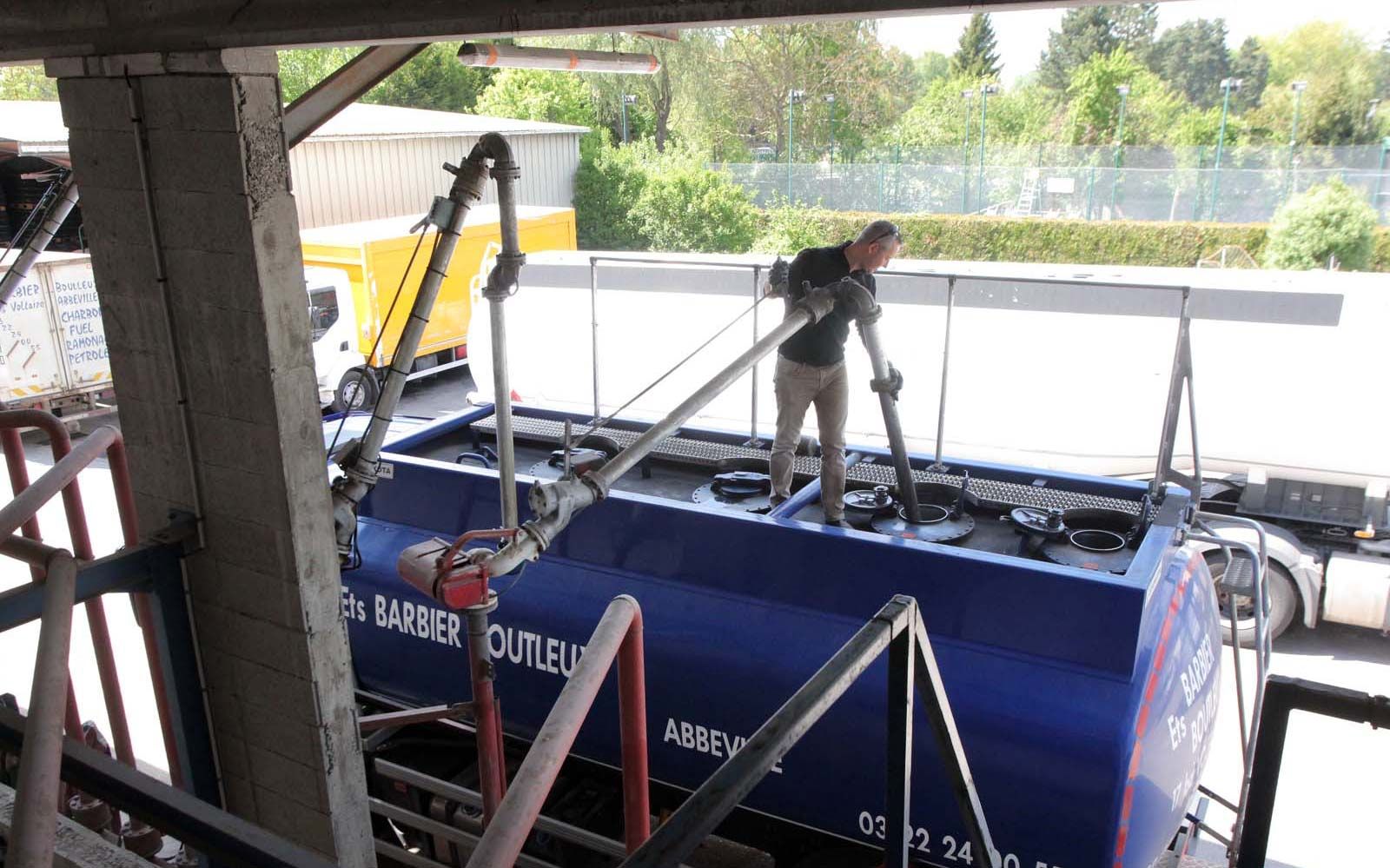 Une personne se tient debout sur un camion-citerne bleu, positionnant un bras de chargement dans une trappe ouverte d'une installation industrielle.