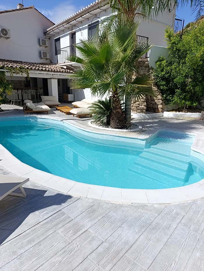 Piscina de agua turquesa rodeada de terraza blanca y un edificio con balcones. Palmera en el centro.