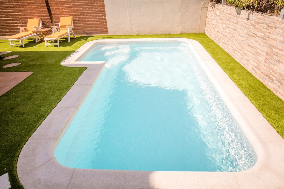 Una piscina rectangular de agua azul claro, rodeada de césped artificial y piedra, con tumbonas cerca.