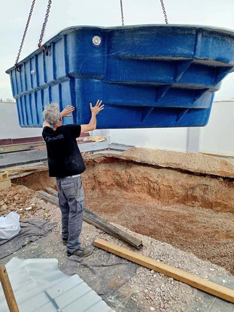 Hombre guiando una carcasa de piscina azul suspendida por una grúa hacia un sitio de piscina excavado.