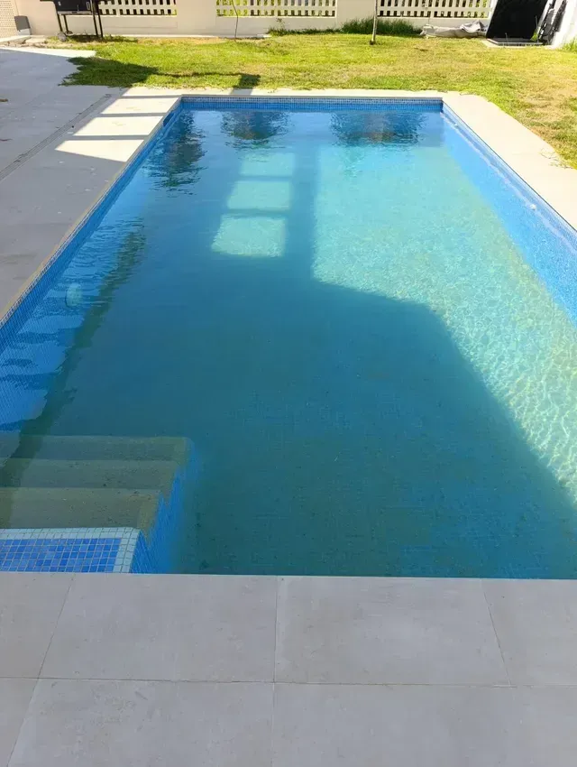 Piscina rectangular de agua azul y escalones, rodeada de piedra blanca y césped verde.