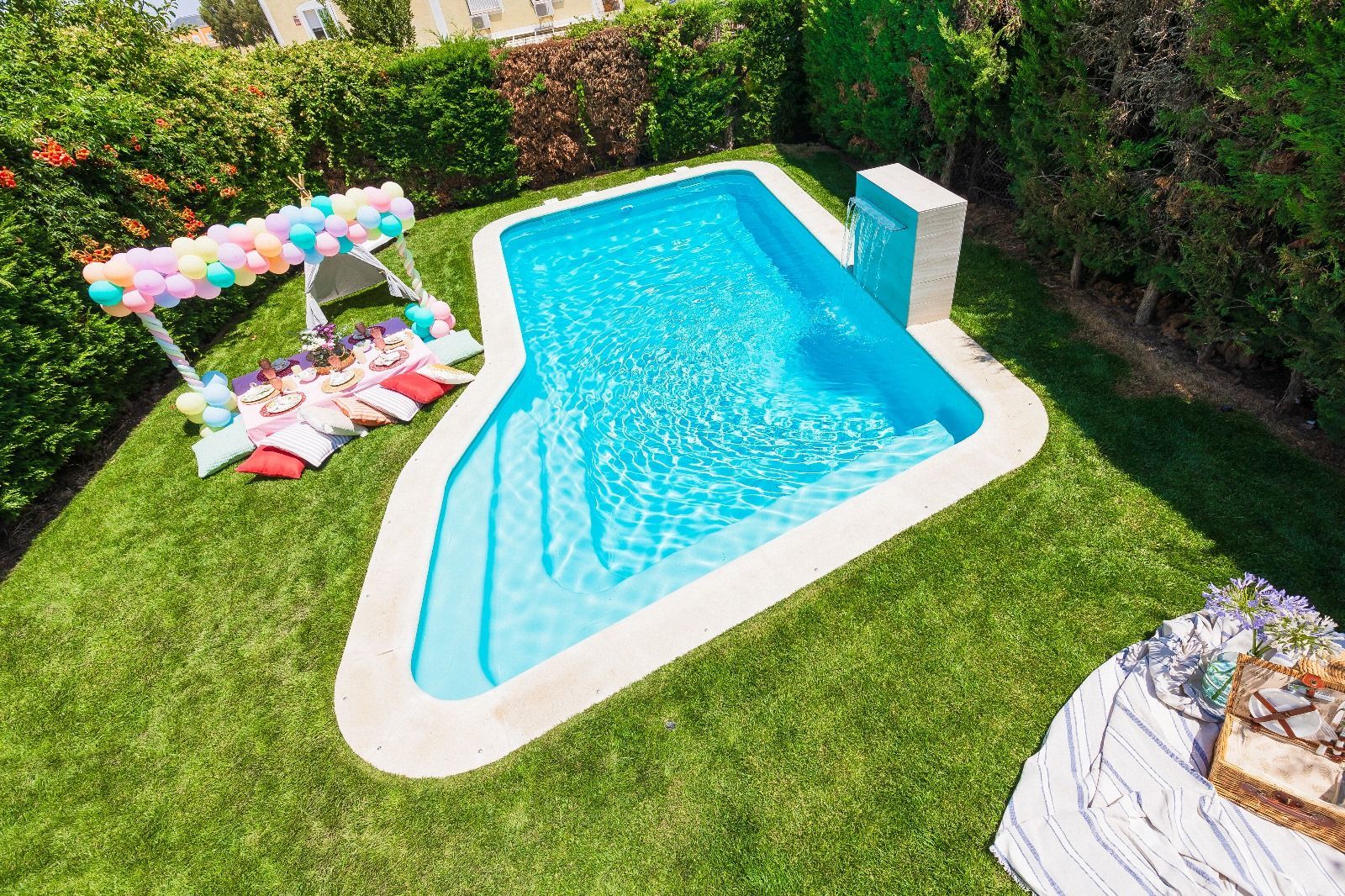 Un patio trasero con una piscina azul. Un área de picnic con globos a la izquierda. Césped verde alrededor.