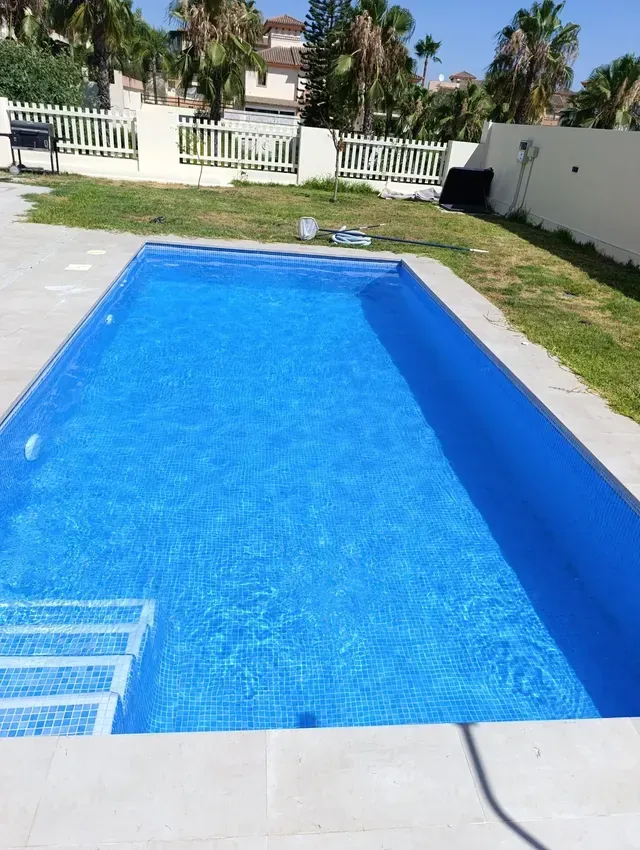 Piscina de azulejos azules en un patio trasero soleado con valla blanca y palmeras.