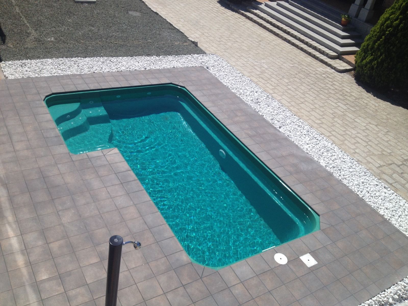 Piscina rectangular de color turquesa con pavimento gris, rodeada de piedras blancas, situada en una soleada zona exterior.