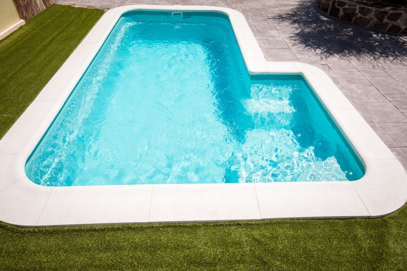 Piscina rectangular con escalones, agua azul, borde blanco y entorno de césped verde.