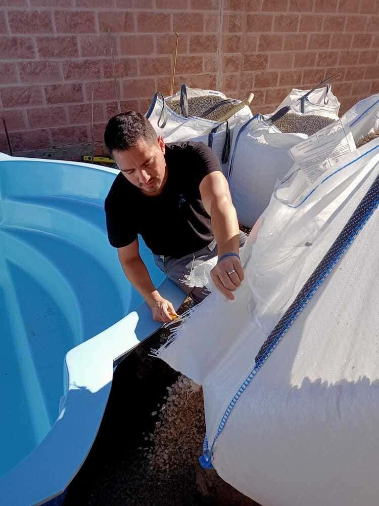 Hombre llenando una piscina azul con grava de grandes bolsas blancas frente a una pared de ladrillos.