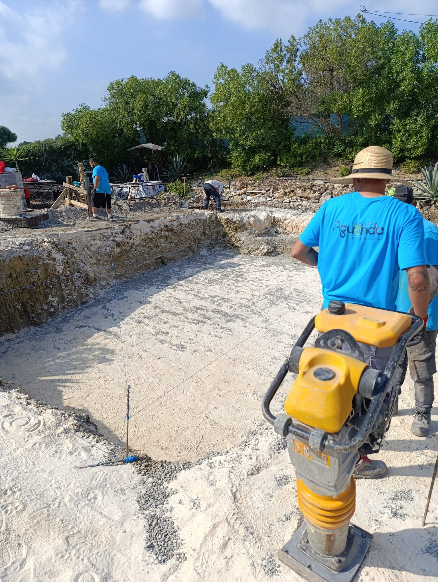 Obreros compactando tierra con una placa vibratoria. Escenario al aire libre con camisas azules, tierra y árboles.