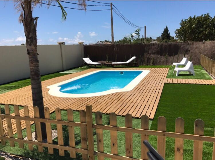 Un área de piscina con una piscina azul, terraza de madera, césped verde y tumbonas blancas bajo un cielo soleado.