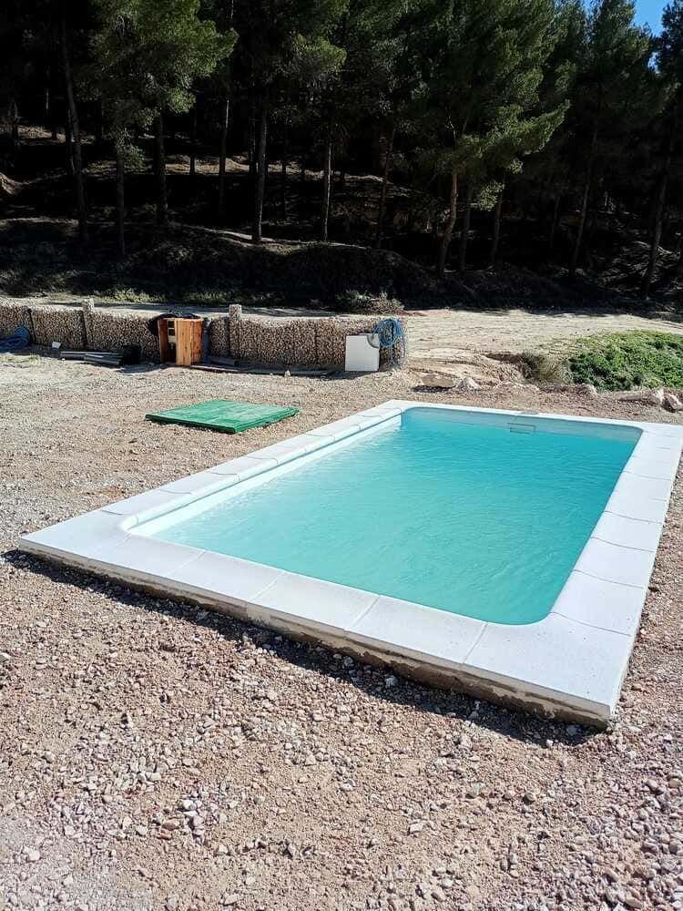 Piscina rectangular de agua azul claro, rodeada de piedra blanca, sobre un terreno de grava con árboles al fondo.