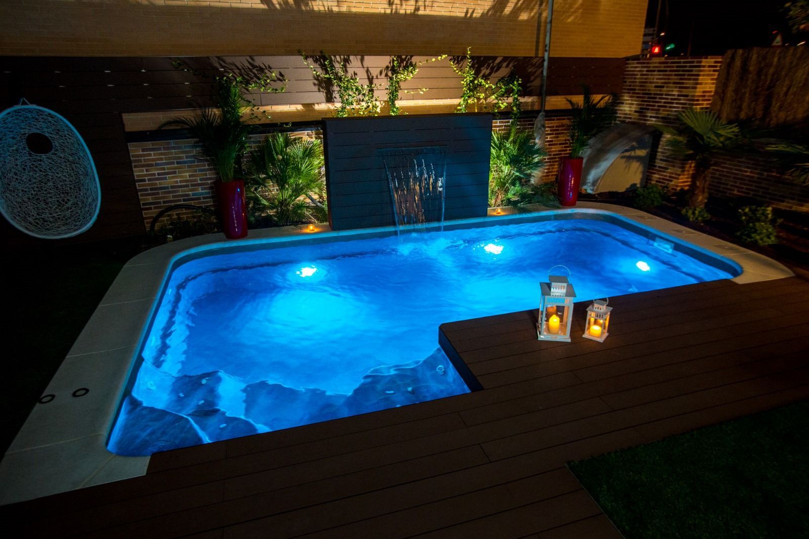 Un pequeño jacuzzi azul iluminado al aire libre por la noche. Terraza con velas, plantas y una fuente.