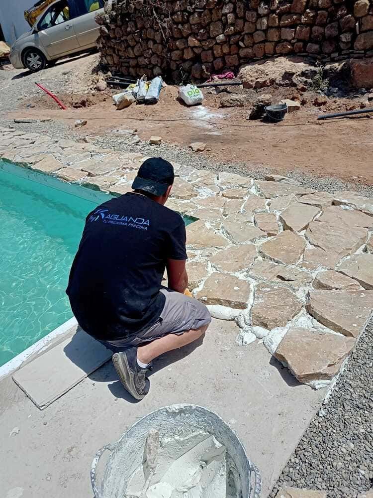 Un hombre coloca un camino de piedra junto a una piscina. Lleva camisa y gorra negras, y el agua es turquesa.