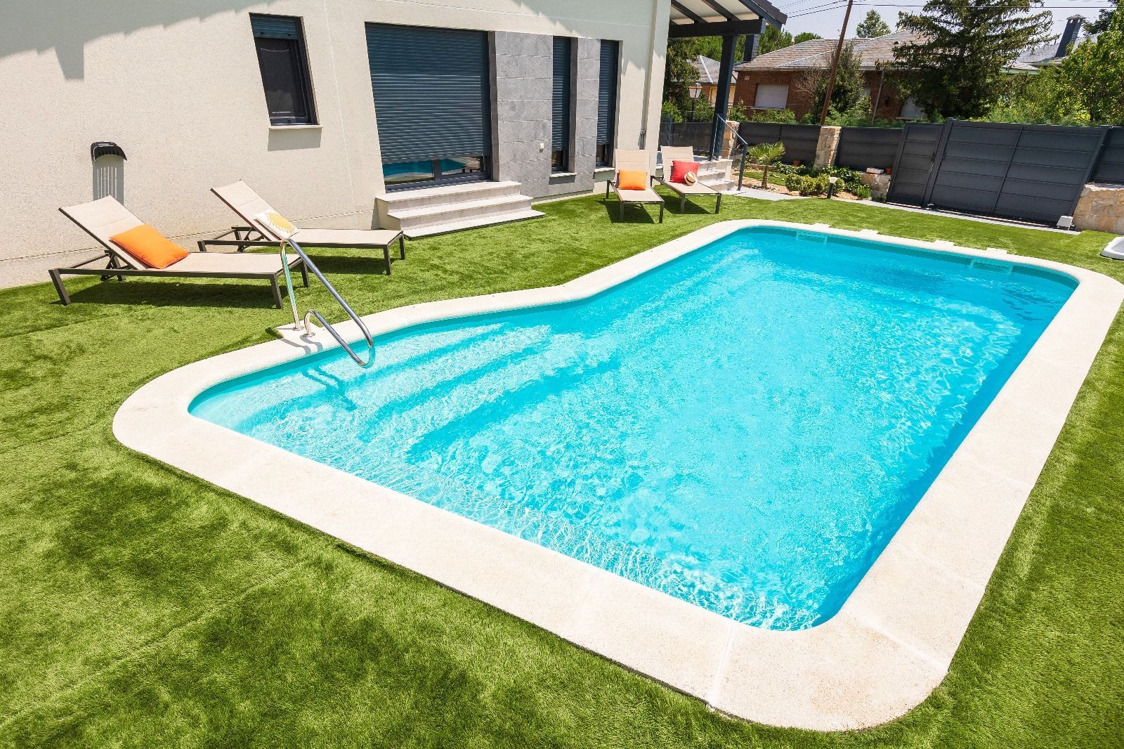 Piscina en el patio trasero con agua turquesa, escalones y césped artificial alrededor. Hay tumbonas cerca.