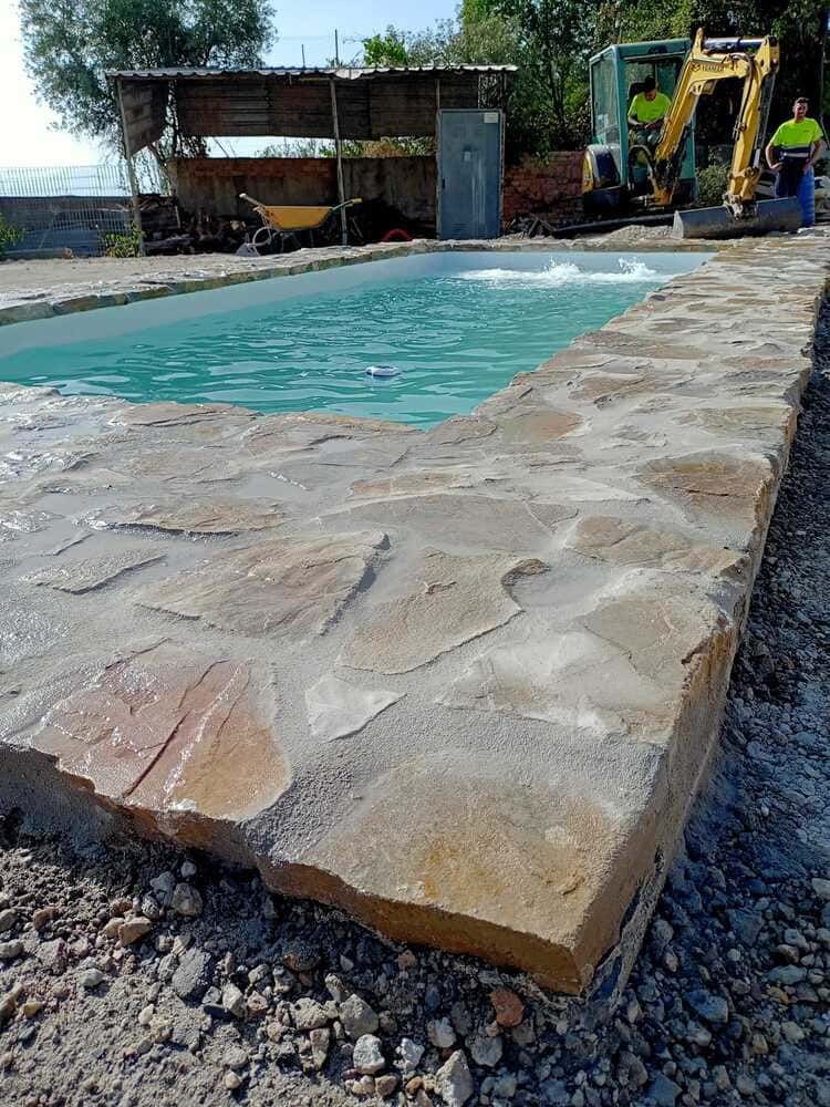 Piscina con borde de piedra en construcción al aire libre, agua turquesa, trabajadores con equipo al fondo.