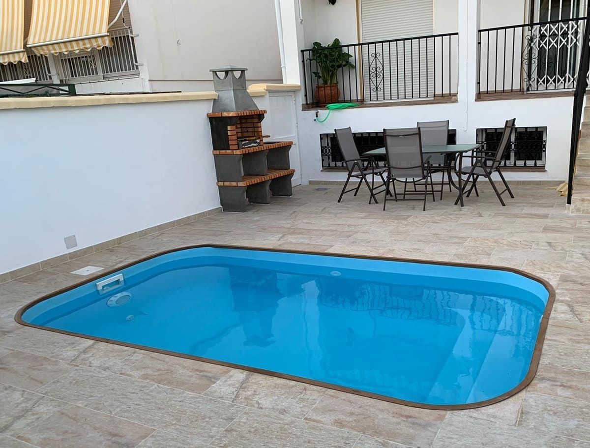 Pequeña piscina rectangular en un patio de mosaico con parrilla, mesa y sillas.