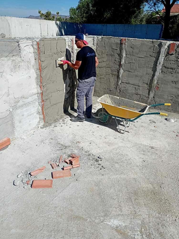 Obrero aplicando mortero a una pared de ladrillos con una llana. Al aire libre, cerca de una carretilla y ladrillos.