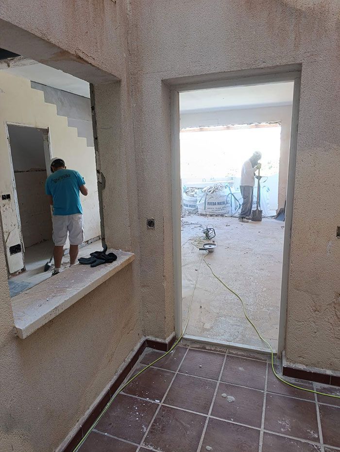 Obra interior con trabajadores cerca de una ventana y una puerta. Paredes rugosas, suelo de baldosas y escaleras visibles.