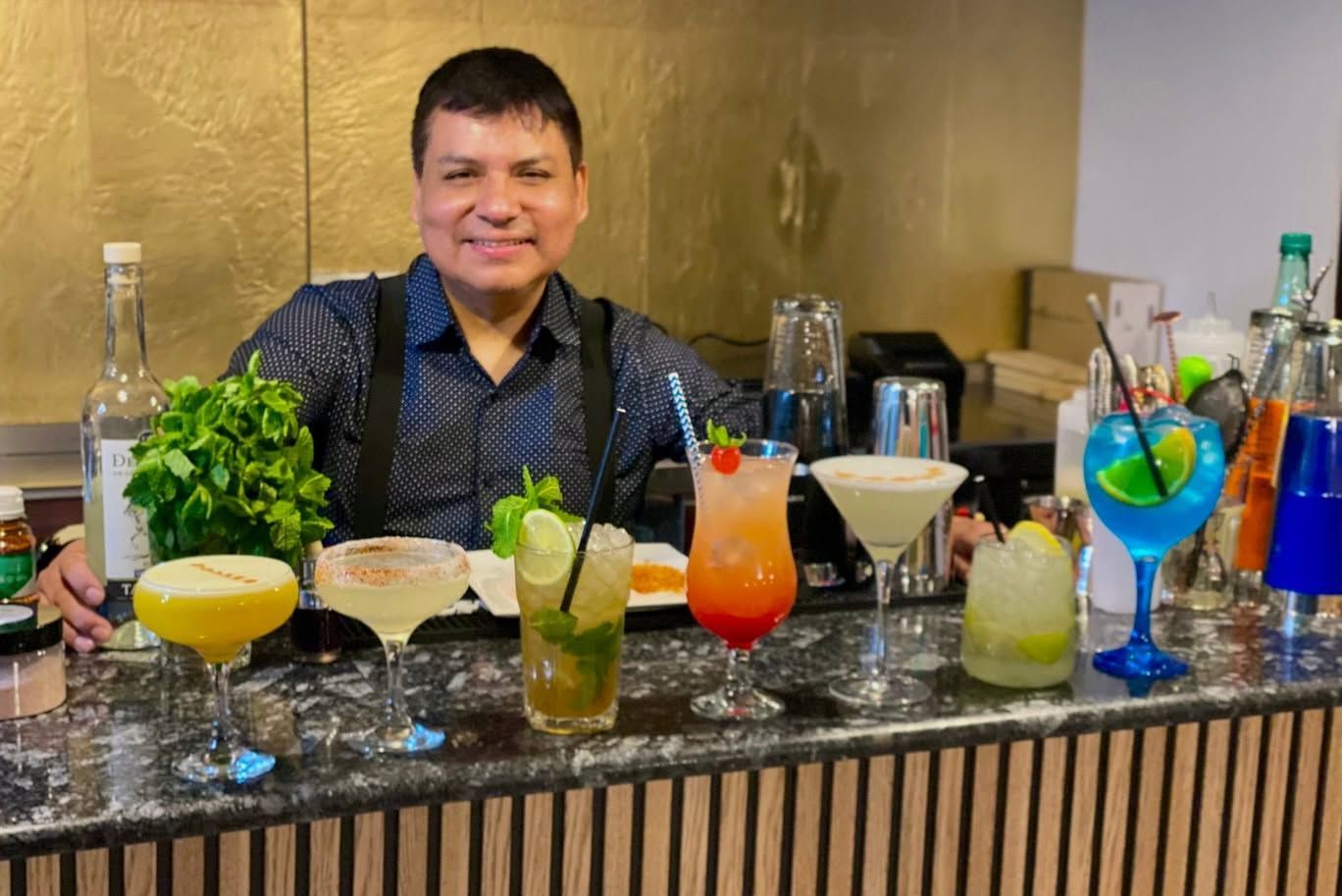 Un hombre de pie detrás de una barra, con cócteles coloridos y botellas de licor sobre una encimera brillante