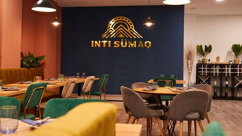 Interior moderno de restaurante con sillas verdes, mesas redondas y un letrero en la pared azul oscuro que dice “INTI SUMAQ”