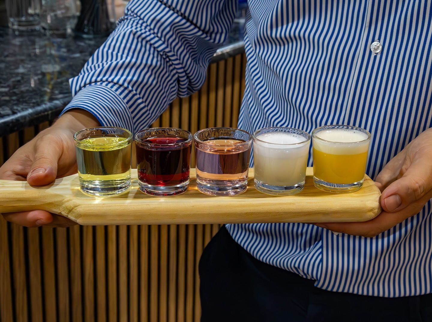 Una persona sosteniendo una bandeja con cinco bebidas de colores en vasos pequeños
